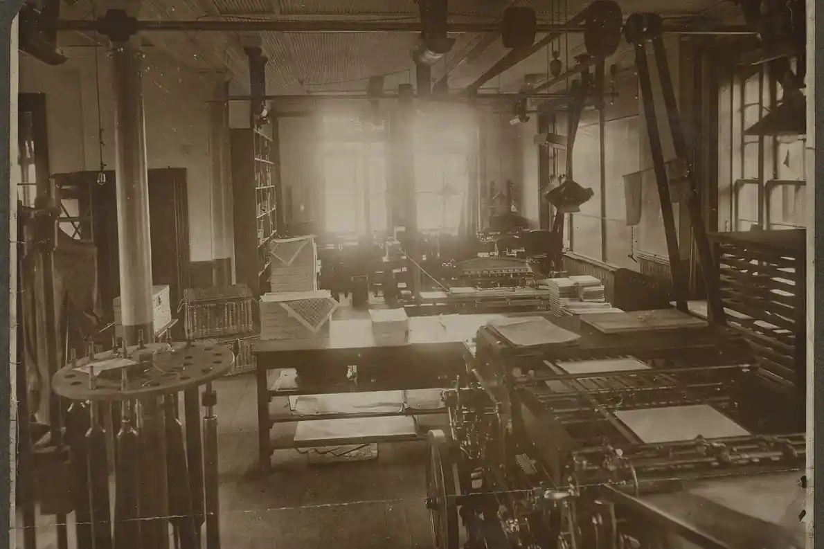 An old printing office seen from the doorway: presses, a long bench stacked with sheets of paper, and tall windows beyond